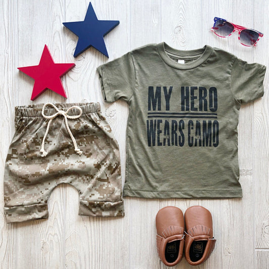 A flat lay features the My Hero Wears Camo • Tee in olive tri-blend with camo shorts, tan moccasins, red-and-blue stars, and sunglasses on a light wood surface. Unisex sizing available.