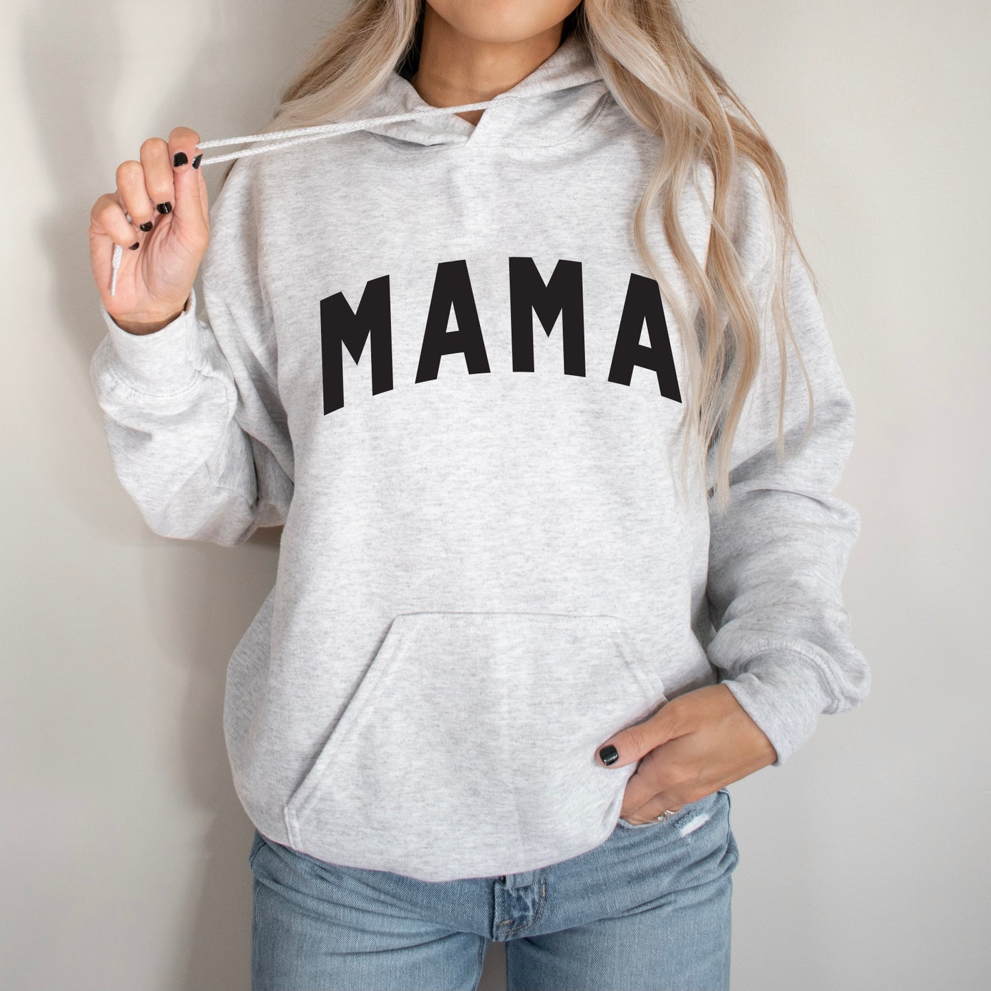 A woman with long, wavy blonde hair wears the MAMA • Ash Hoodie—a light gray, pill-resistant fleece sweatshirt with MAMA in bold black letters. She holds one hoodie string and has her other hand in her jeans pocket.