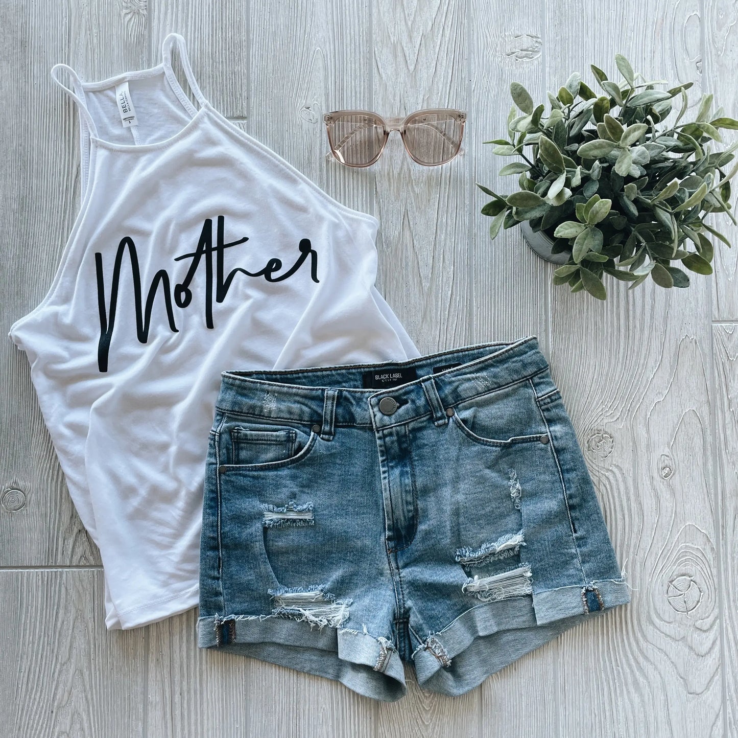 A flat lay features the Mother • High Neck Tank—a white high-neck women’s top with Mother in black—paired with distressed denim shorts, rose gold sunglasses, and a potted plant on light wood, highlighting its relaxed drapey fit.