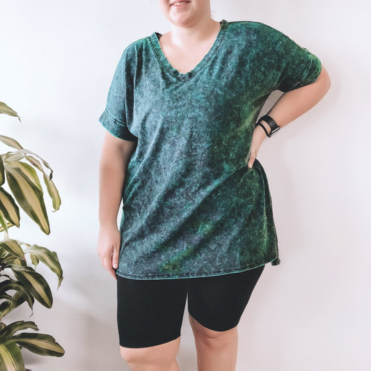 A person wearing a loose, green tie-dye V-neck T-shirt and black Brushed Biker Shorts stands with one hand on their hip next to a green leafy plant, against a plain white background.