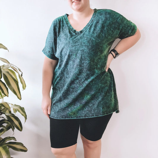A person wearing a loose, green tie-dye V-neck T-shirt and black Brushed Biker Shorts stands with one hand on their hip next to a green leafy plant, against a plain white background.