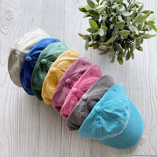 Seven Mama Life Hat • NEW Colors! canvas caps—beige, blue, green, yellow, pink, gray, and turquoise—some with vintage-inspired embroidery, are lined up neatly on a light wooden surface beside a leafy potted plant.