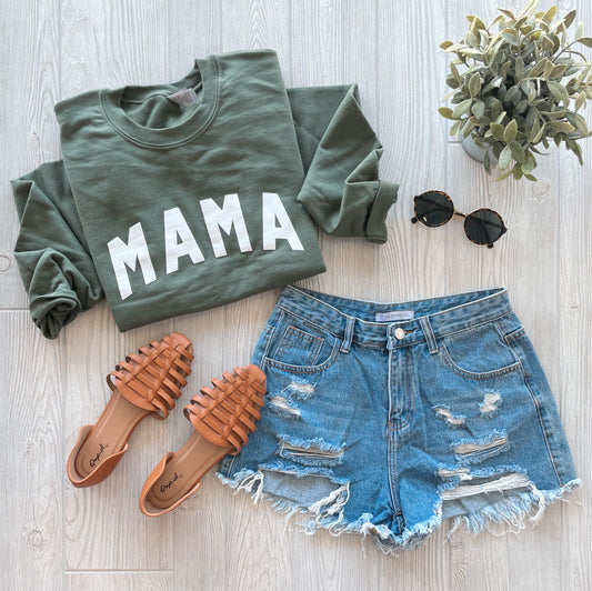 Flat lay of the MAMA • Military Green Pullover, styled with ripped denim shorts, tan woven sandals, round sunglasses, and a small potted plant on a light wood floor.