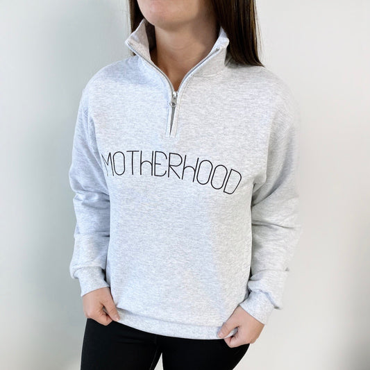 A woman wears the Motherhood • Quarter Zip Pullover in light gray, featuring MOTHERHOOD in bold black letters across the chest, standing against a plain white background.