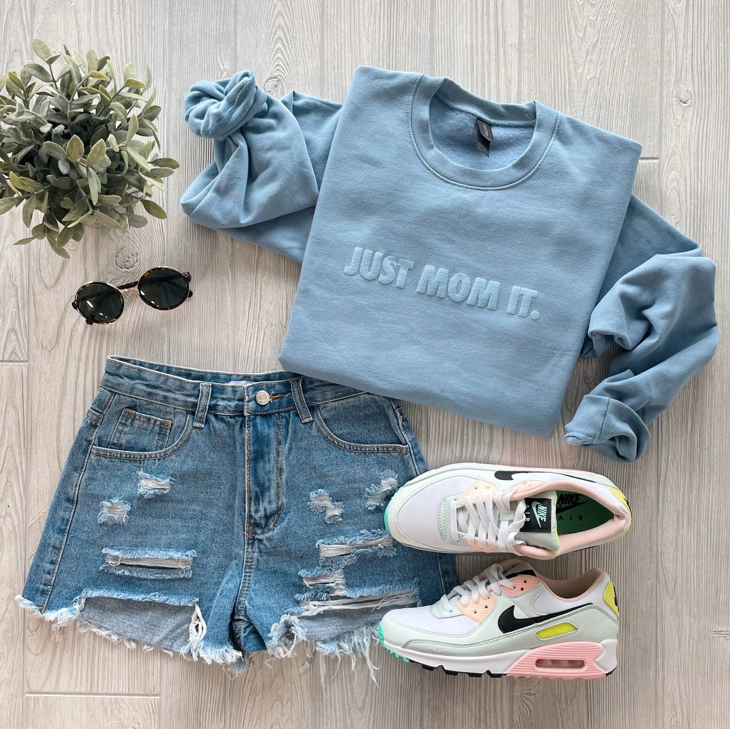 Flat lay: JUST MOM IT • Steel Blue Pullover (unisex, ring spun cotton), distressed denim shorts, white pastel-accented sneakers, black sunglasses, and a small green plant on a light wood floor.