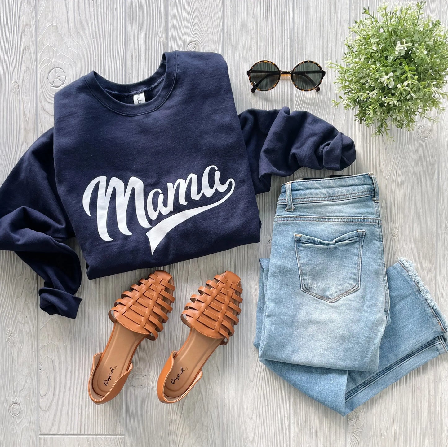 A flat lay of the Mama Sporty • Navy Pullover, a unisex-fit sweatshirt with “Mama” in white script, styled with light blue jeans, brown woven sandals, round sunglasses, and a green potted plant on a light wood floor.