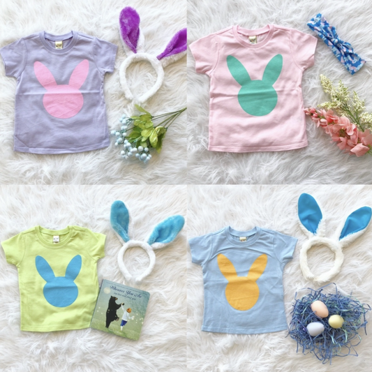 Four Original Bunny • Kids Tees in Kavio size specs are paired with matching bunny ear headbands and spring props, displayed on a white fluffy background in a charming flat lay arrangement.