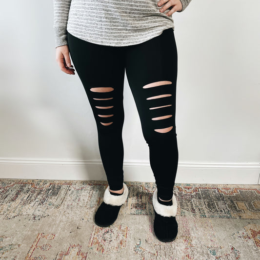 Wearing New Shredded Leggings, a person in a striped long-sleeve shirt and black slippers with white trim stands on a patterned rug before a white wall, highlighting stylish casual athleisure wear.