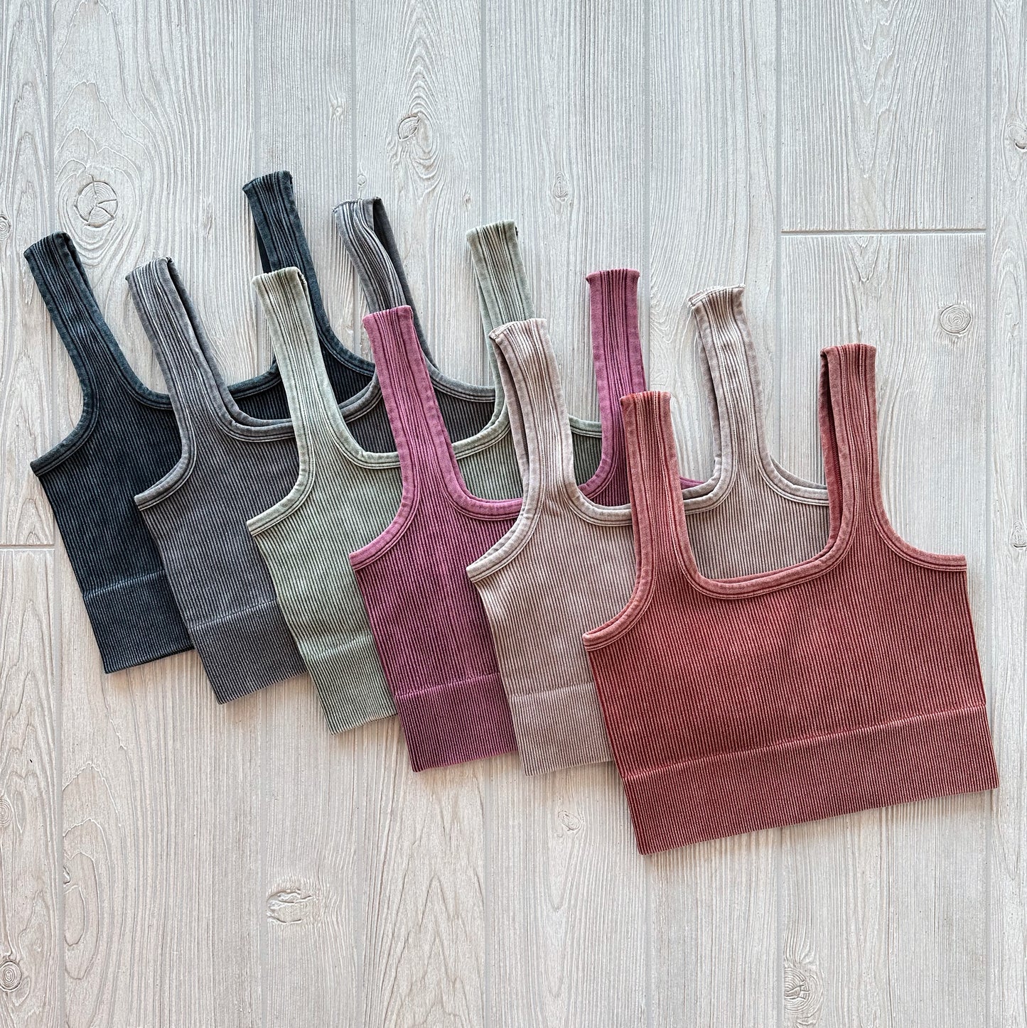 Seven Iris Crop tops from the Denver Collection in muted colors—dark gray, charcoal, olive, taupe, mauve, dusty rose, and rust—are lined up on a light wooden floor to highlight their comfy, stretchy ribbed fabric.