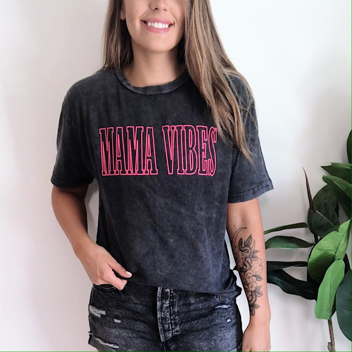 A smiling woman with long hair and a forearm tattoo stands indoors by a green potted plant, wearing the MAMA VIBES • Rocker Tee in black with bold pink lettering. Made from soft Ringspun Combed Cotton for comfort and style.