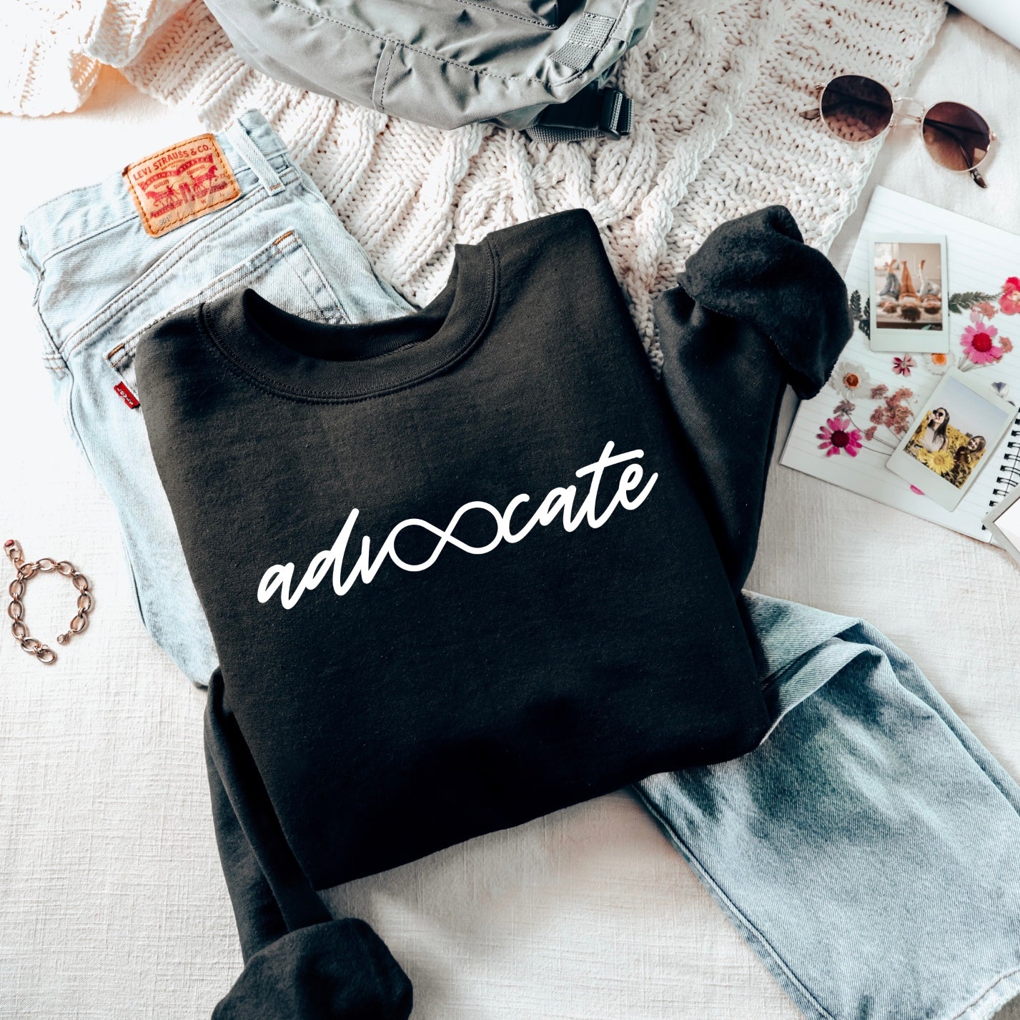 The Advocate • Black Pullover, a unisex sweatshirt in black with “advocate” and an infinity symbol, is folded on jeans. Soft cotton/polyester blend. Shown with a knit sweater, sunglasses, bracelet, flower photos, notebook, and gray backpack.