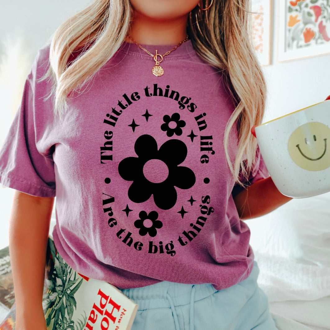 A blonde woman with wavy hair holds a mug and magazine, wearing The Little Things • Comfort Colors Tee in purple featuring black floral designs and the phrase “The little things in life are the big things.”.