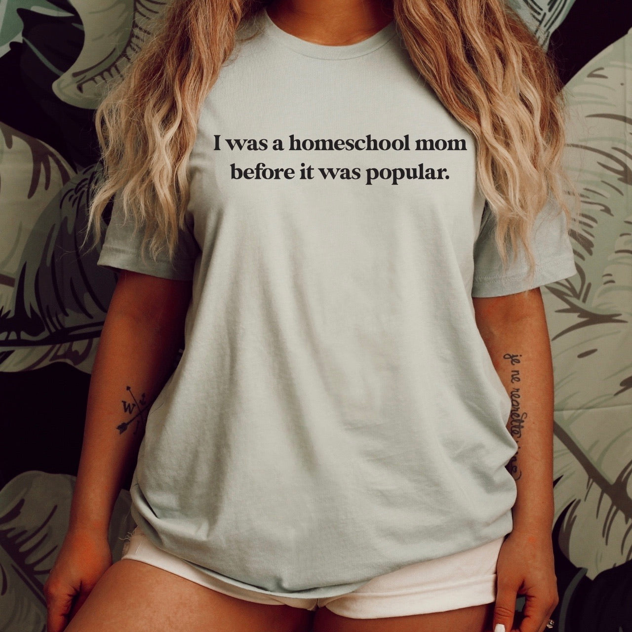 A woman with long, wavy hair wears the Homeschool Mom • Tee, a light gray Bella Canvas shirt with "I was a homeschool mom before it was popular" printed on it. She stands in front of a leafy patterned background, her face not shown.