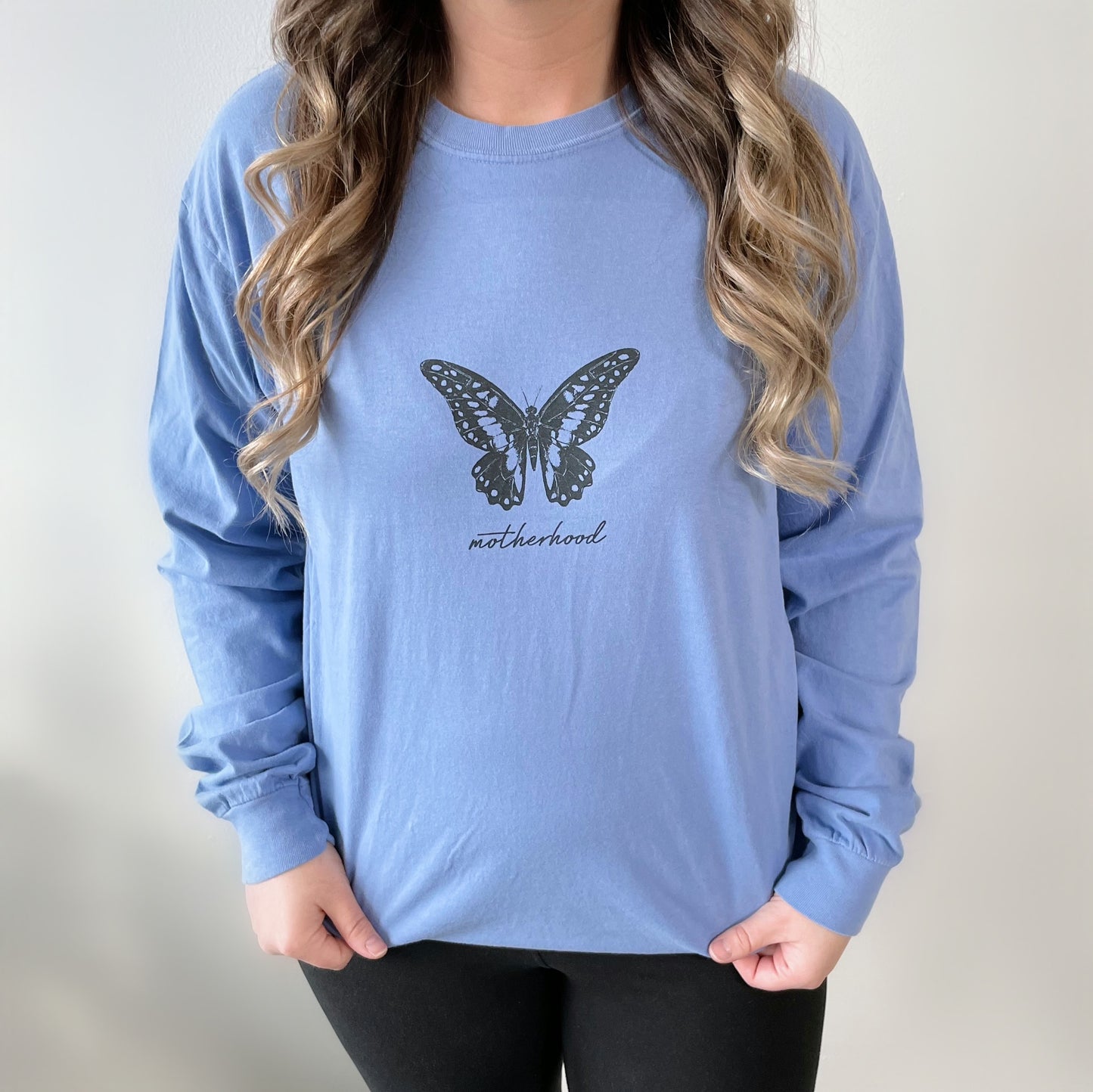 A woman wearing the Butterfly • Periwinkle Long Sleeve, a soft ring-spun cotton crew neck shirt featuring a black butterfly graphic and motherhood text. She stands against a plain light background with long, wavy hair.