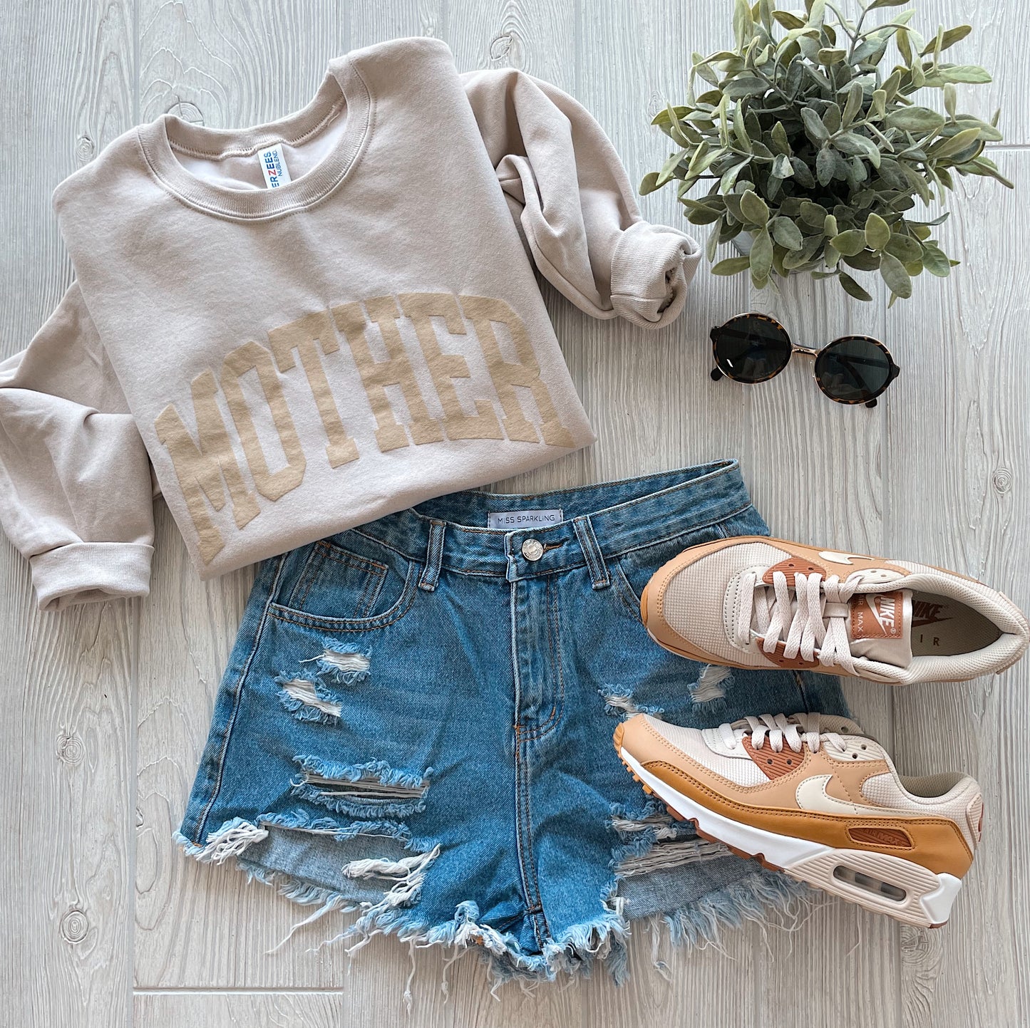 A MOTHER Tonal Sand Pullover, distressed denim shorts, beige and peach Nike sneakers, black sunglasses, and a small potted plant are arranged on light wood. This unisex sweatshirt is crafted from a soft cotton-polyester blend.