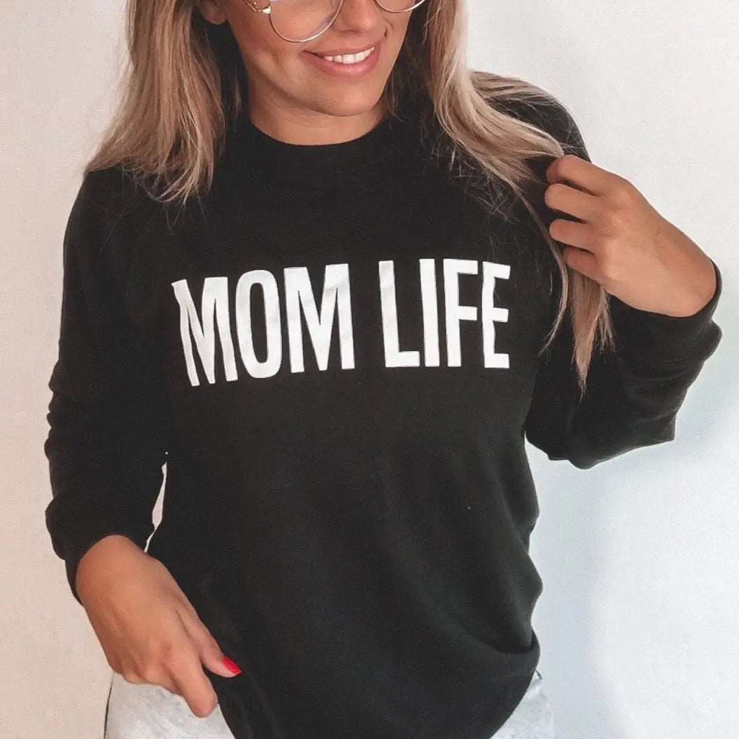A woman with long hair and glasses smiles while wearing the MOM LIFE • Black Sweatshirt, a unisex fit pullover with “MOM LIFE” boldly printed in white letters.