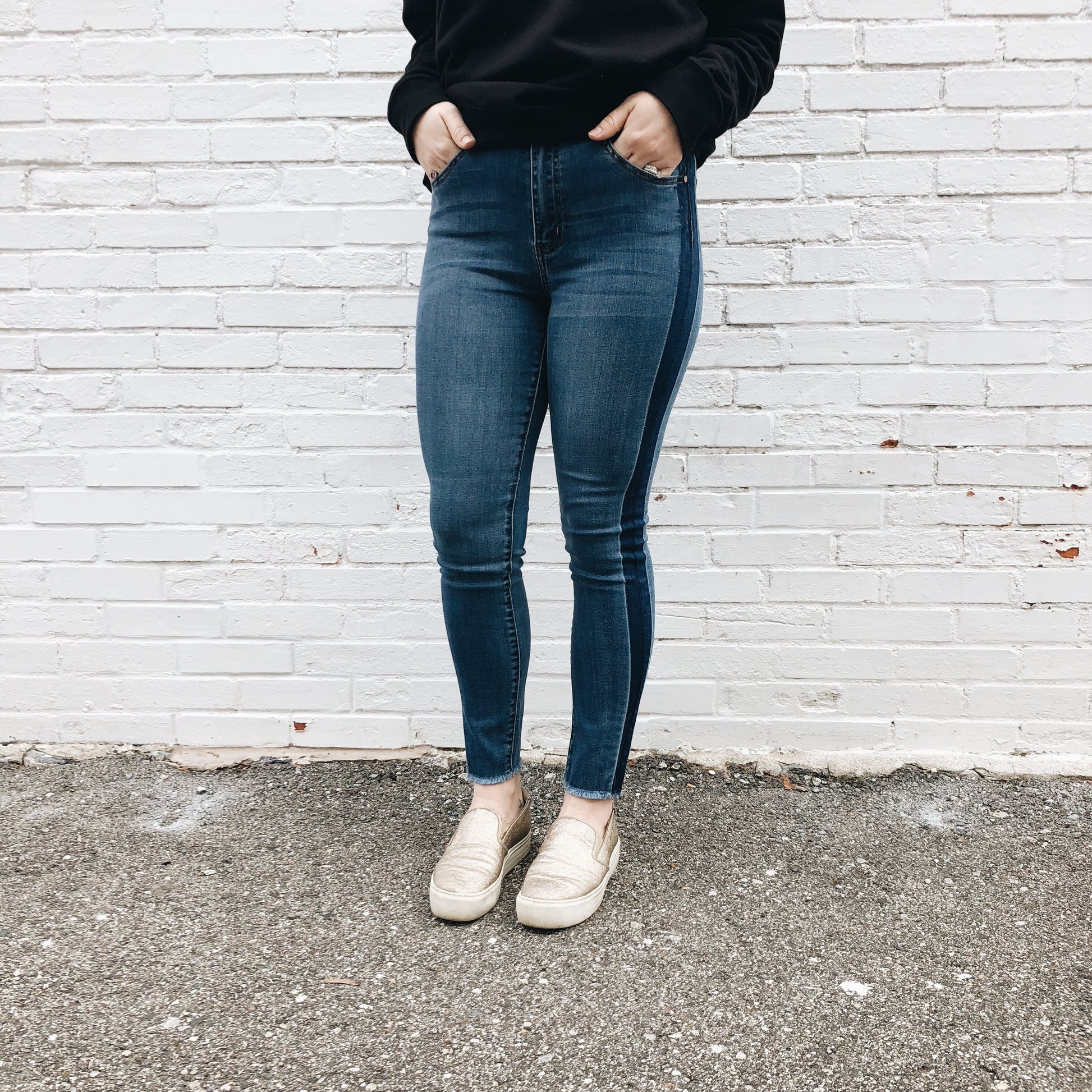 Standing against a white brick wall, the person wears Alie Jeggings SIZE 0 & 3 that fit true to size, a black sweatshirt, and beige slip-on shoes. The photo is cropped at the shoulders, keeping their face out of frame.