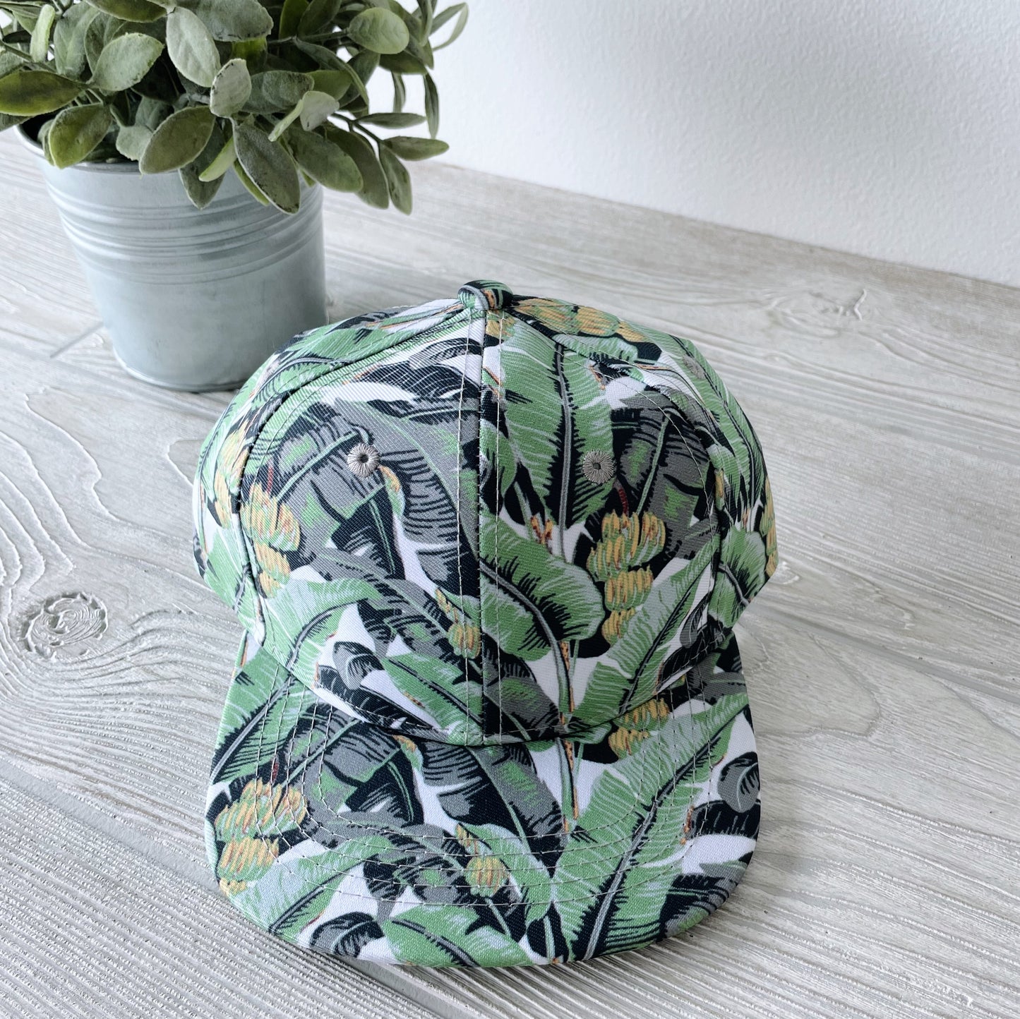 The Kids Snapback • Easton, featuring a green tropical leaf and yellow flower print, rests on a light wood surface beside a potted plant with small, rounded green leaves.