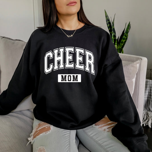 A woman sits on a couch wearing the Cheer Mom • Black Pullover, a unisex sweatshirt with bold white CHEER MOM lettering, paired with ripped light gray jeans. A green plant appears in the background.