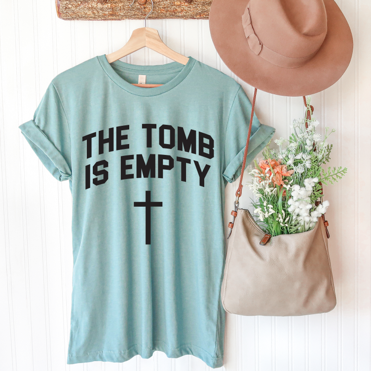 The Resurrection • Dusty Blue Tee, featuring THE TOMB IS EMPTY and a cross, hangs on a wooden hanger beside a tan hat and beige flower-filled bag on a white wall. Unisex sizing ensures it’s perfect for everyone.