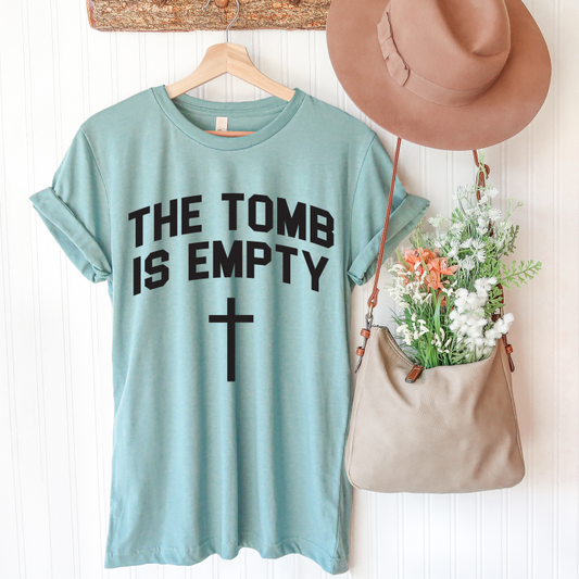 The Resurrection • Dusty Blue Tee, featuring THE TOMB IS EMPTY and a cross, hangs on a wooden hanger beside a tan hat and beige flower-filled bag on a white wall. Unisex sizing ensures it’s perfect for everyone.