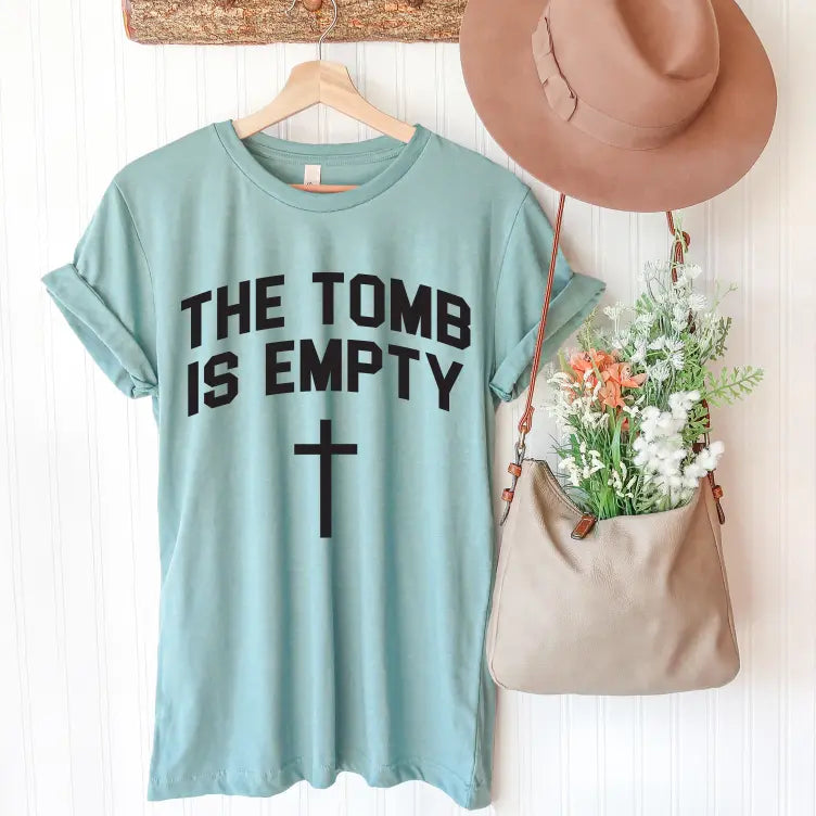 The Resurrection • Dusty Blue Tee, featuring THE TOMB IS EMPTY and a cross, hangs on a wooden hanger beside a tan hat and beige flower-filled bag on a white wall. Unisex sizing ensures it’s perfect for everyone.