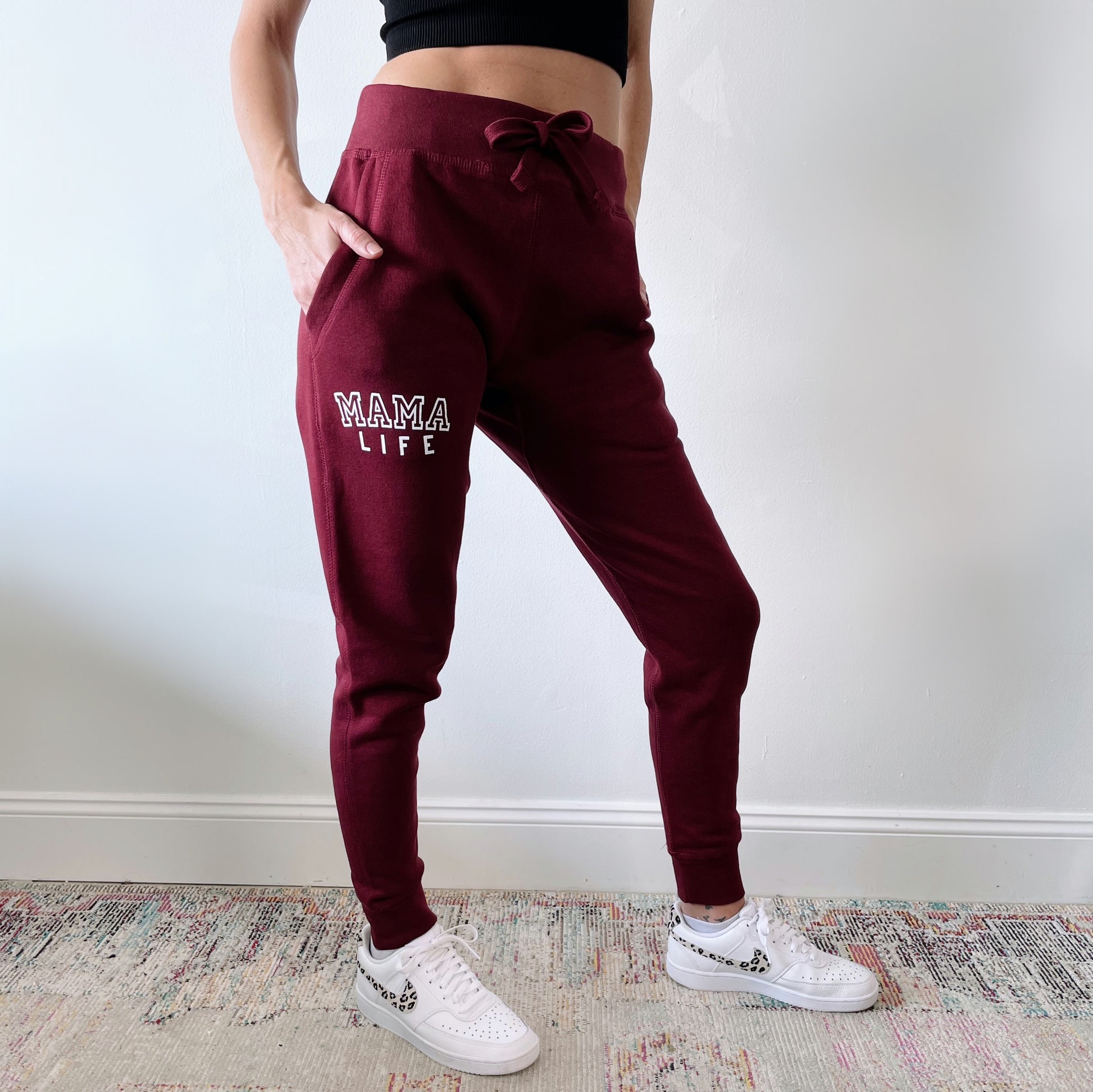 Someone wears the Mama Life • Maroon Joggers featuring MAMA LIFE on one leg, paired with a black crop top and white sneakers, standing indoors on a colorful rug. Ideal for cozy, stylish loungewear.