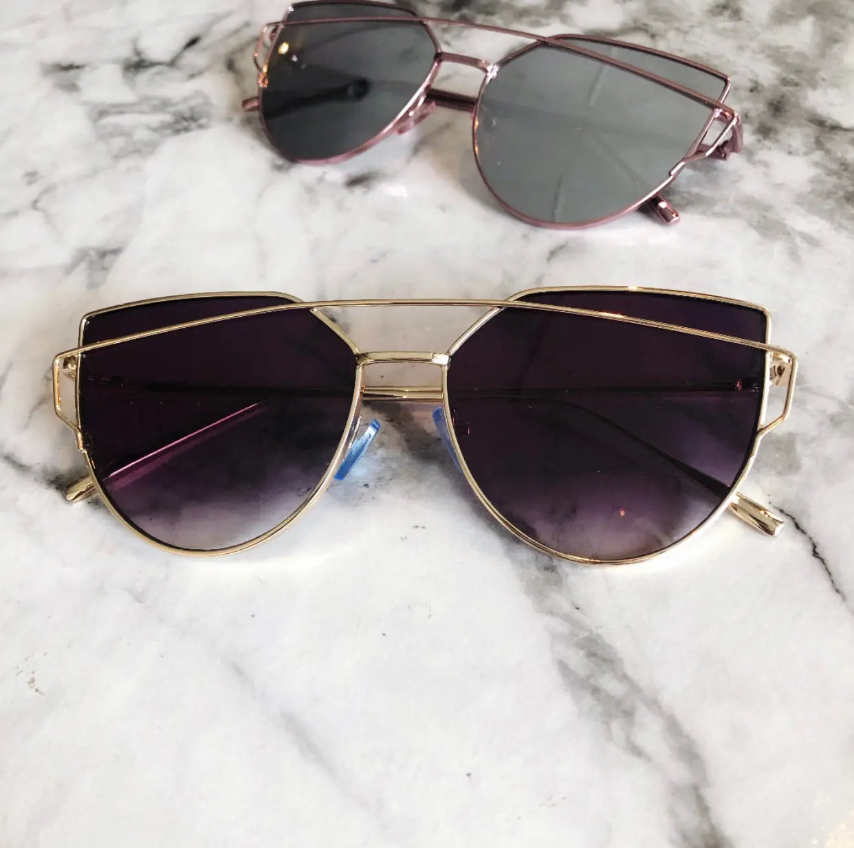 Two pairs of Claire adult sunglasses in more colors, featuring gold frames and UV-protected tinted lenses, rest on white marble. One has dark purple lenses in the foreground; the other is gray—both ready to ship.