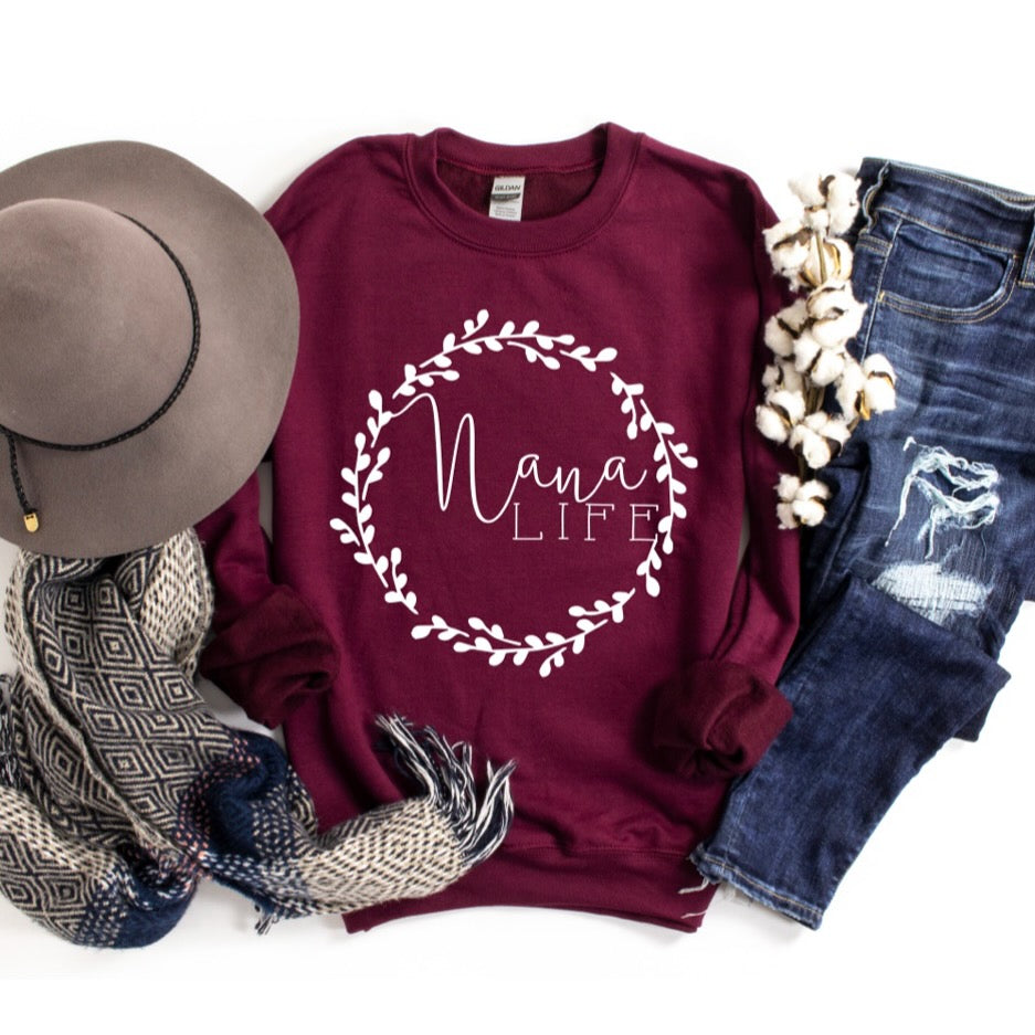 Flat lay of the Nana Wreath • Tee or Pullover—a burgundy unisex fit with “Nana Life” and a white wreath. Styled with distressed jeans, gray hat, scarf, and cotton branch—perfect for fans of charming grandma designs.