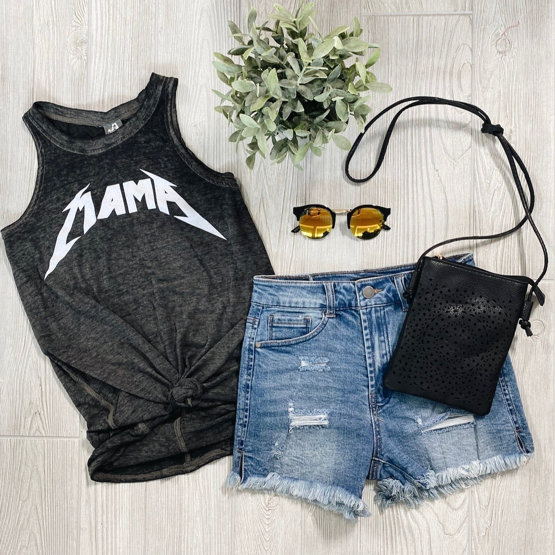 Flat lay of a casual outfit: the black sleeveless Metal MAMA • Grunge Tank in burnout jersey, distressed denim shorts, yellow-lens sunglasses, a black crossbody bag, and a potted green plant on light wood flooring.