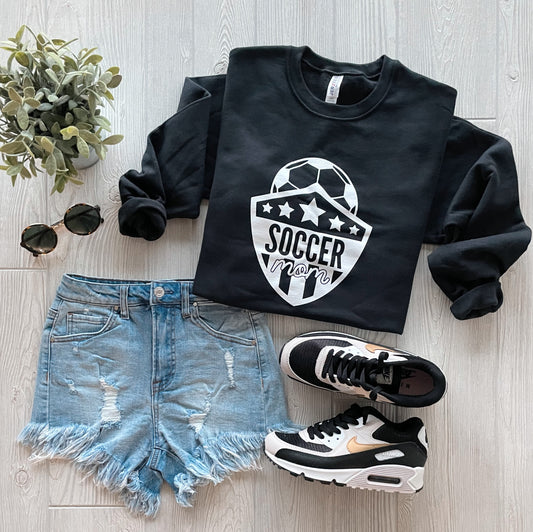 Flat lay of the Soccer Mom • Black Pullover styled with distressed denim shorts, black and white sneakers, round sunglasses, and a small green plant on a light wood floor.