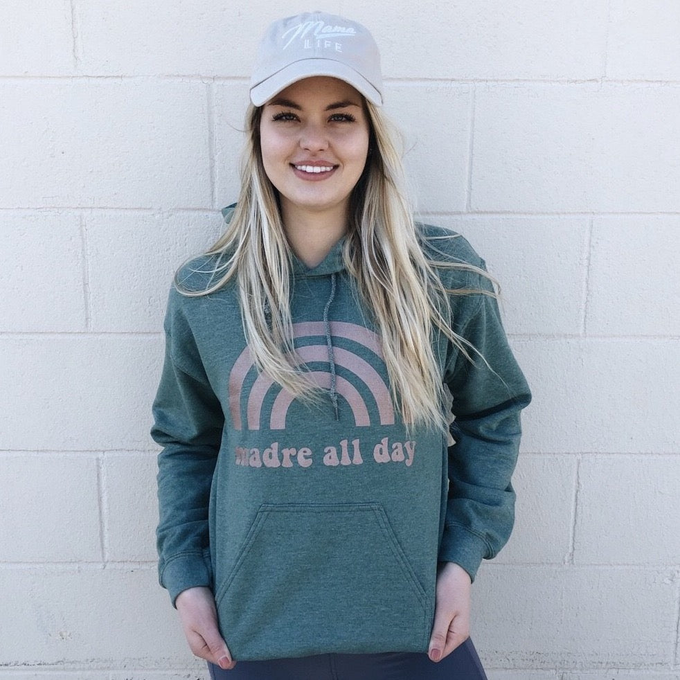 A woman smiles against a light brick wall, wearing a light-colored Mama Life cap and the Madre All Day • Vintage Forest Hoodie with a rainbow print.