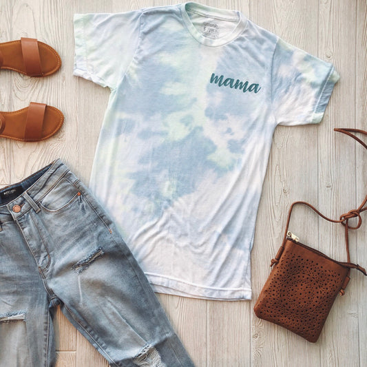 Flat lay of a casual outfit: Mama Script • Green Tie-Dye tee, made from a soft polyester cotton blend, paired with light wash ripped jeans, brown sandals, and a cutout brown crossbody bag on a wooden background.