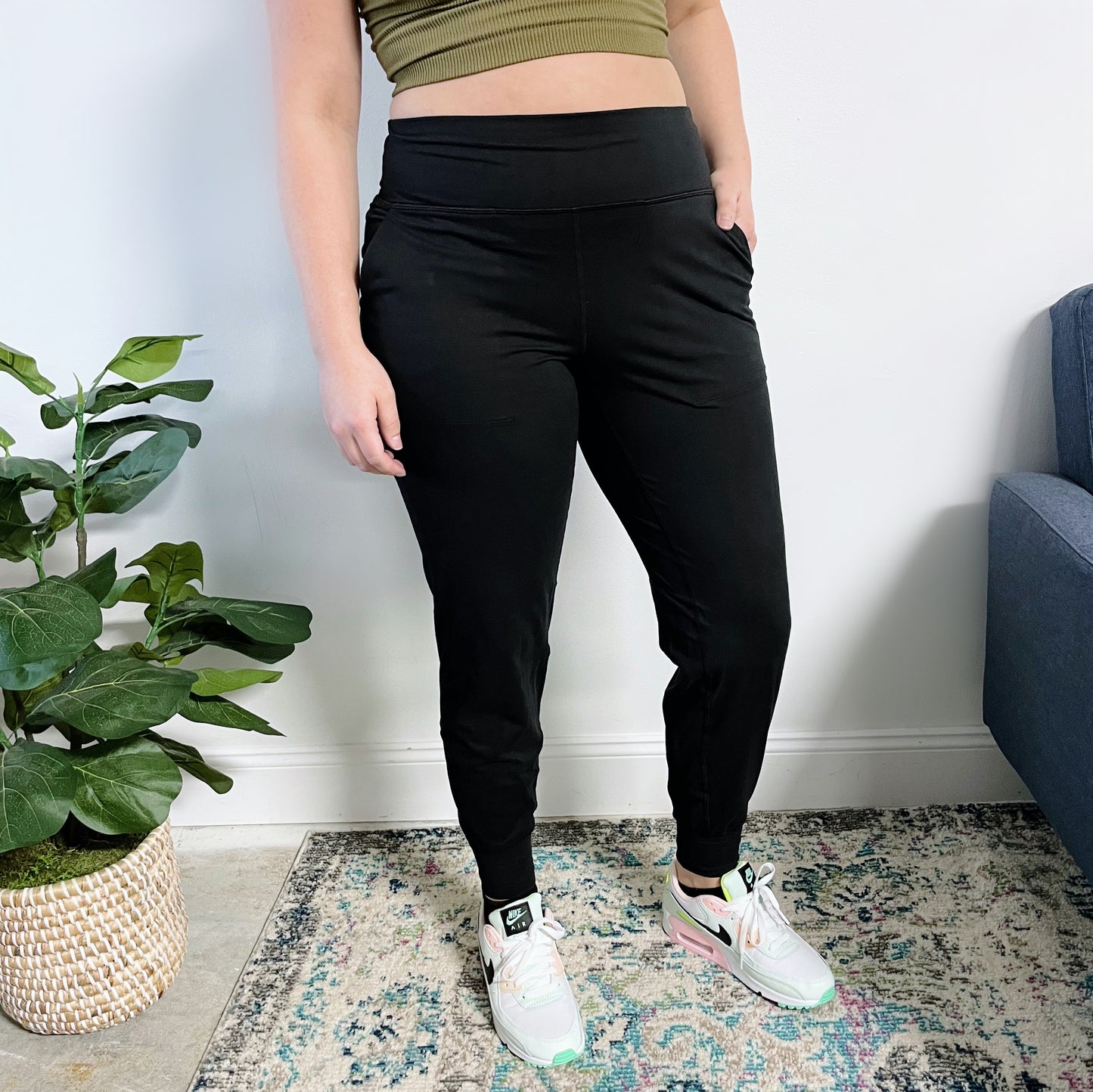 A person stands indoors on a patterned rug beside a potted plant and blue chair, wearing white sneakers, a green crop top, and the Lexi Yoga Pant in Black with their left hand in their pocket.
