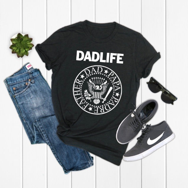 Flat lay of a black Dad Life • Band Tee unisex t-shirt, blue jeans, gray Nike sneakers, black sunglasses, and a small potted succulent arranged on a white wooden surface.