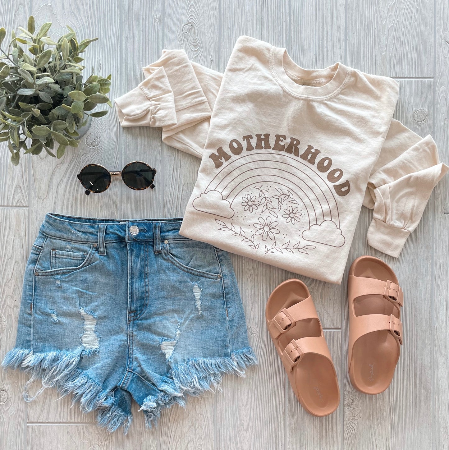 Flat lay of a Motherhood • Cream Long Sleeve with a rainbow graphic, paired with distressed denim shorts, tan slide sandals, black round sunglasses, and a small potted plant on a light wooden floor.