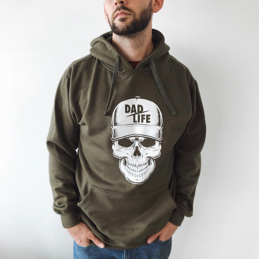 A man with a beard stands with his hands in his pockets, wearing the Dad Life Skeleton • Olive Hoodie. The cotton/polyester fleece features a skull graphic with a DAD LIFE cap, shown against a plain white background.