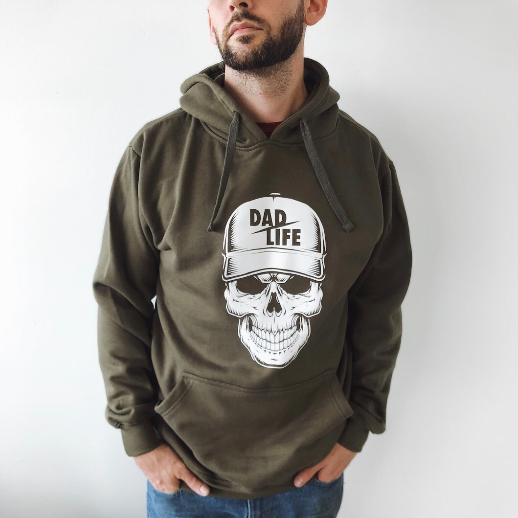 A man with a beard stands with his hands in his pockets, wearing the Dad Life Skeleton • Olive Hoodie. The cotton/polyester fleece features a skull graphic with a DAD LIFE cap, shown against a plain white background.