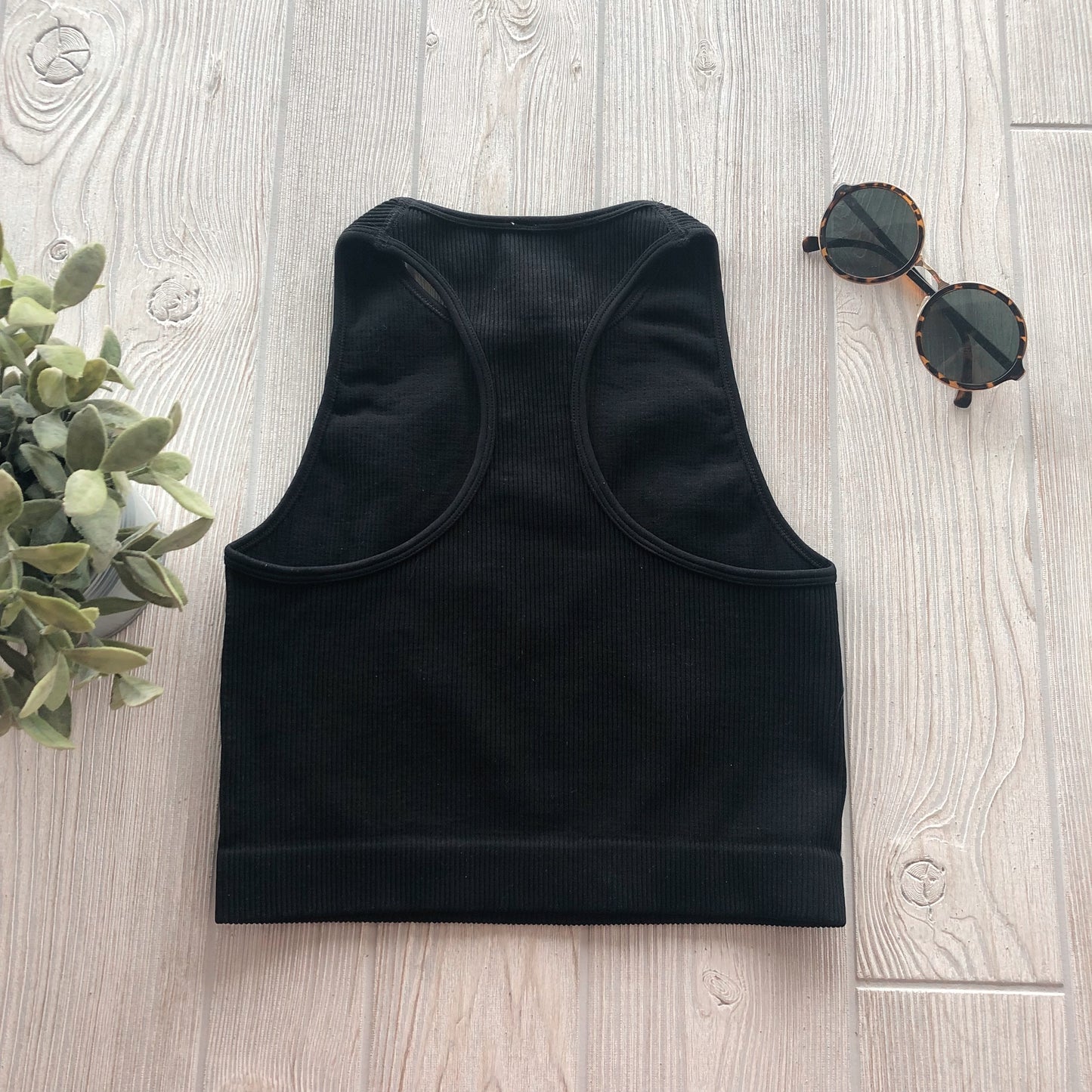 The Sophia High-Neck Crop • Black is laid flat on a light wood surface, beside round tortoiseshell sunglasses and a small green plant.