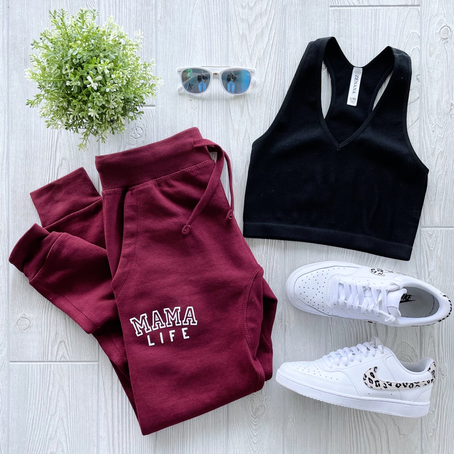 A flat lay on a light wooden floor features Mama Life • Maroon Joggers, a black sports bra, white sneakers with spotted laces, mirrored sunglasses, and a small potted plant.