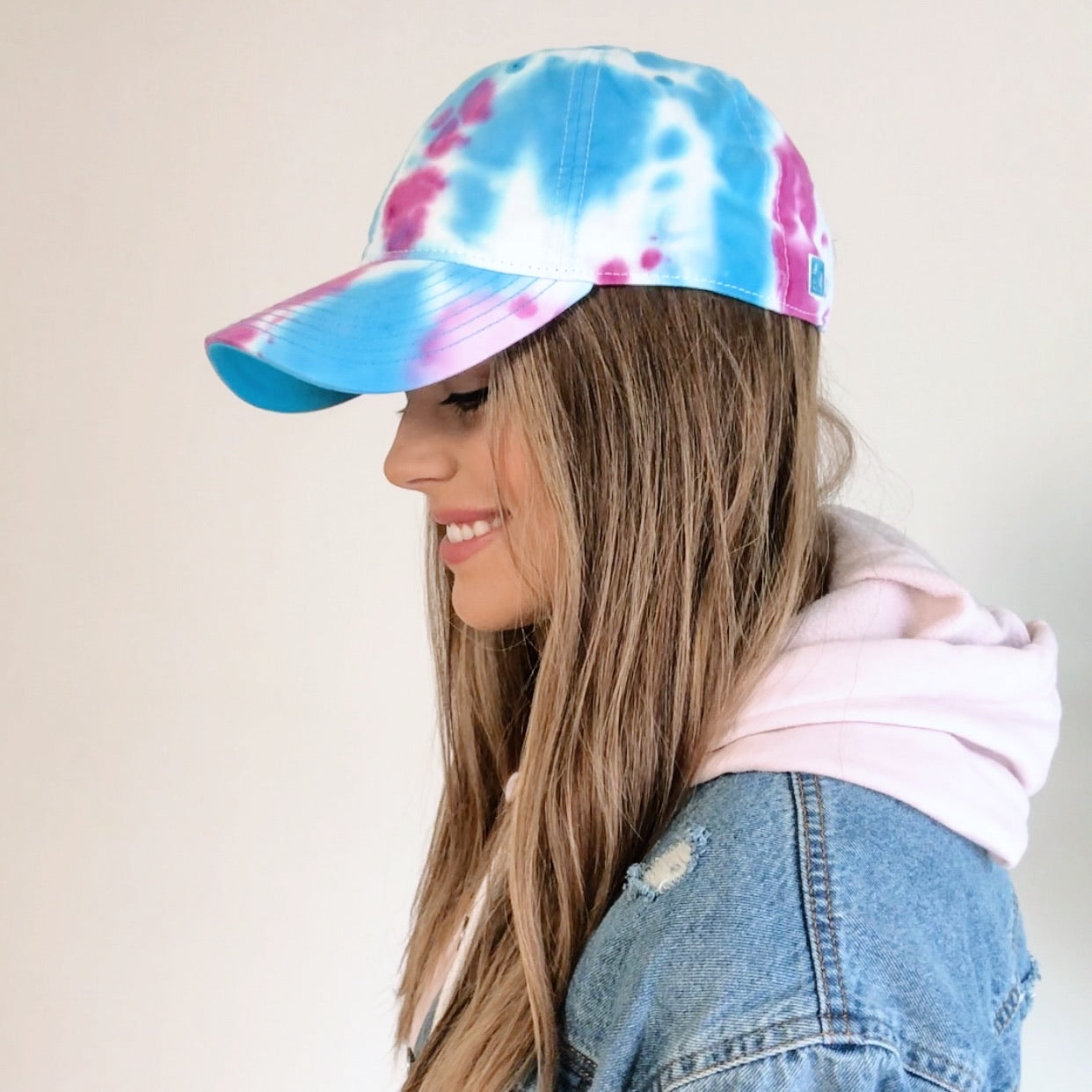 A woman with long, light brown hair smiles while wearing the Tie Dye Hat • Jelly Bean in blue, pink, and white, paired with a pale pink hoodie and denim jacket, facing left against a plain background.