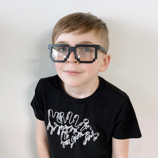 A young boy with light brown hair wears Minecraft Glasses • Kids—pixelated block-style frames—and a black Killing It shirt, standing against a plain white wall. Please check measurements before purchase; all sales are final.