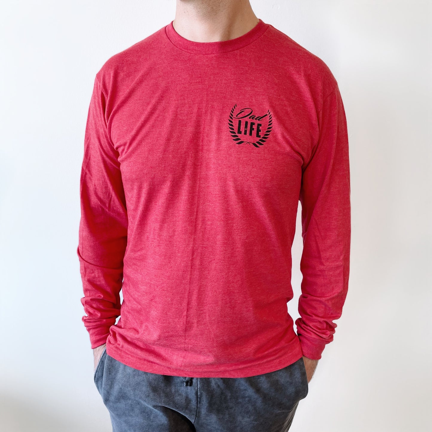 A person wears the Dad Life Crest • Long Sleeve, a red unisex shirt with a Dad Life and laurel graphic on the upper left chest, made from soft ring-spun cotton, standing against a plain white background with hands in their pockets.