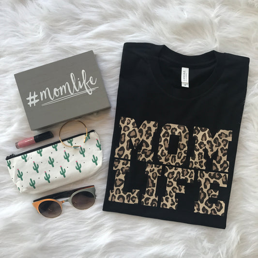 Flat lay of the Mom Life • Cheetah Tee, a black unisex Bella Canvas t-shirt with bold cheetah print MOM LIFE, plus #momlife gray box, cactus pouch, sunglasses, bracelet, and pink lip gloss on a white furry background. Everyday comfort.
