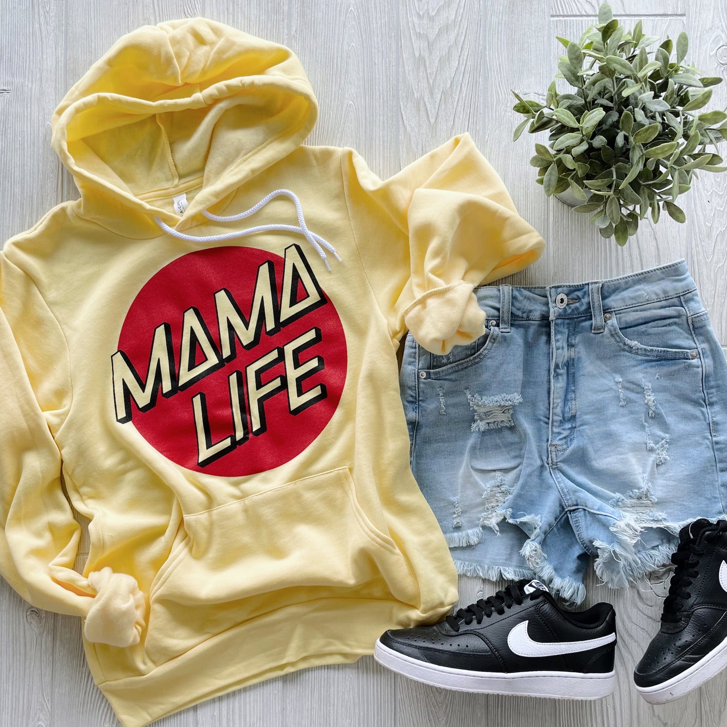 The Mama Life • Yellow Skater Hoodie is styled with distressed denim shorts, black Nike sneakers, and a small green potted plant, set on a light wood background.