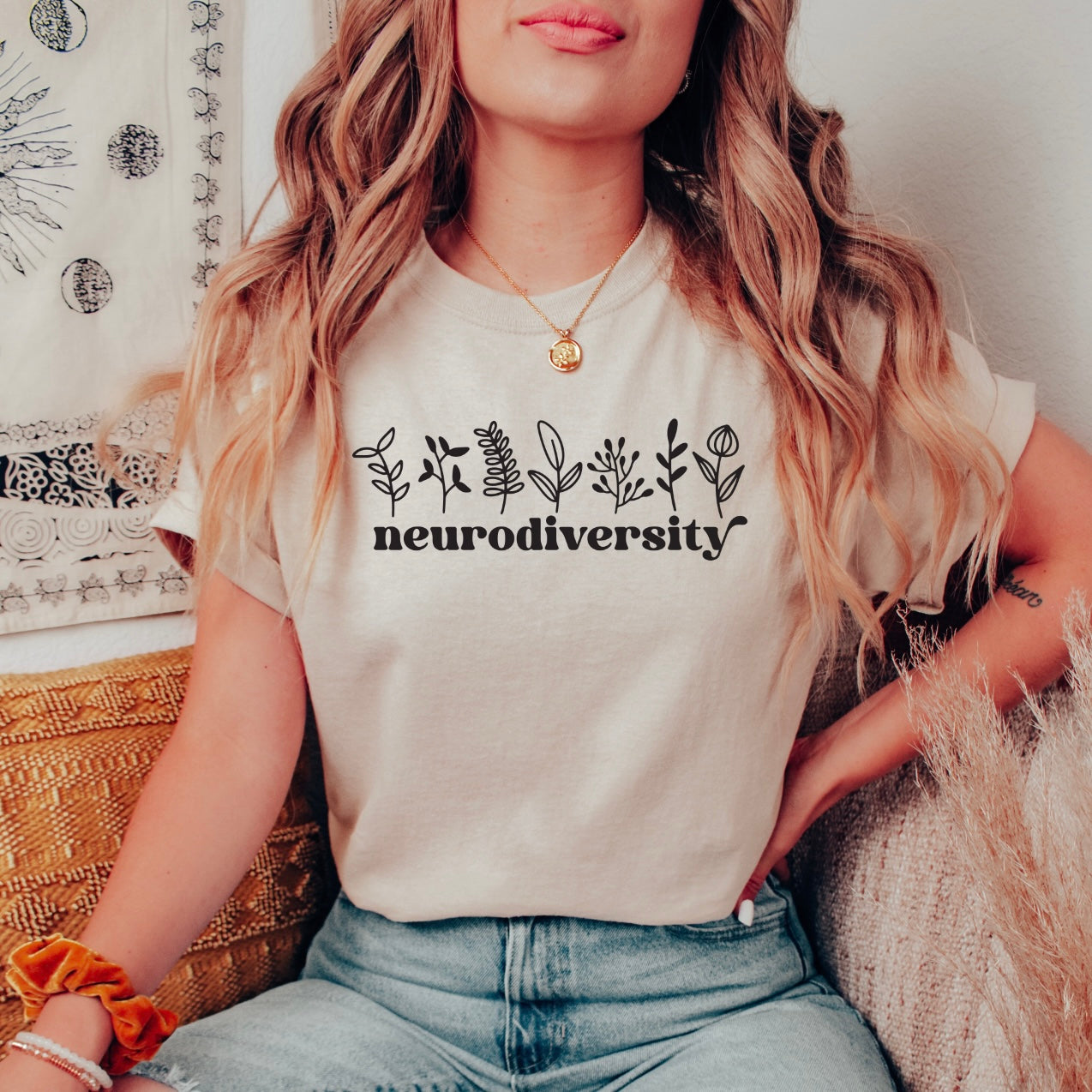 A woman with wavy hair and a gold necklace sits on a woven chair wearing the Neurodiversity • Cream Tee, featuring plant illustrations above the text. The retail fit stands out against her light blue jeans and a patterned tapestry backdrop.