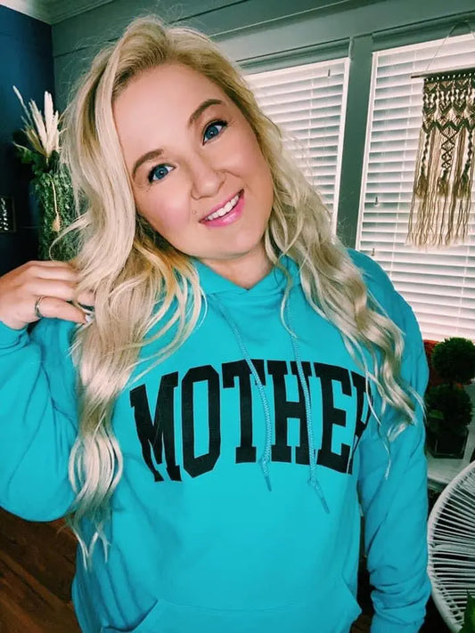 A smiling woman with long blonde hair wears the MOTHER • Aqua Hoodie, a soft fleece unisex fit. She stands indoors by windows with white blinds and decorative plants.