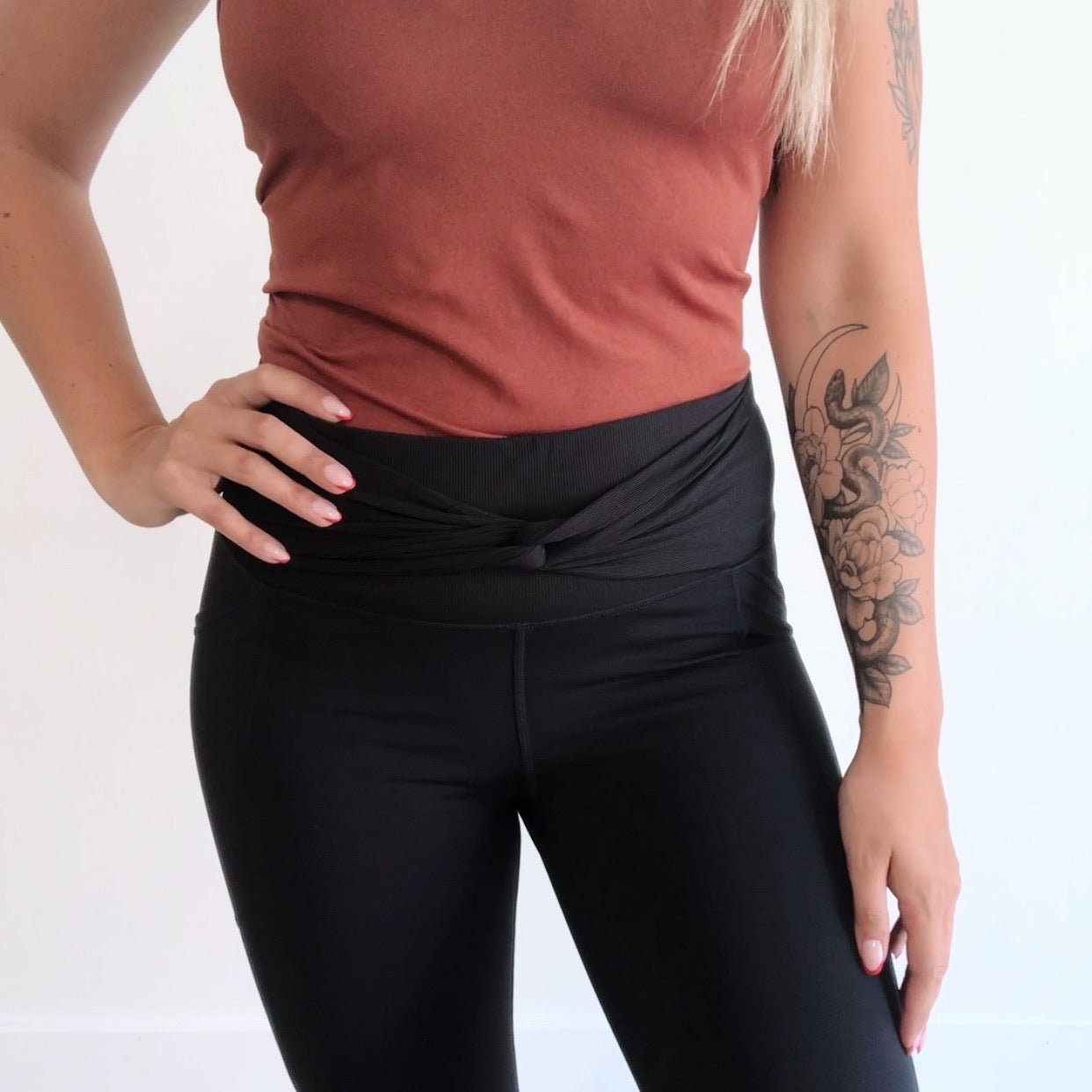 A woman with a floral forearm tattoo stands with one hand on her hip, wearing a reddish-brown sleeveless top and black Twist Accent Highwaist Legging • 2869.