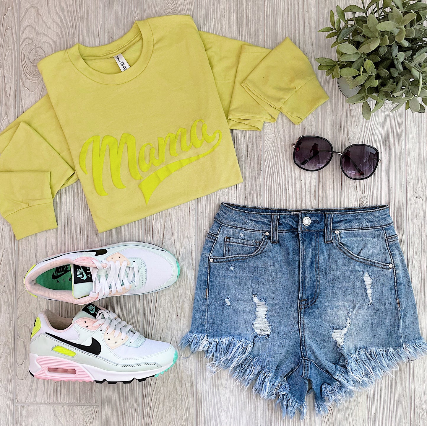 Flat lay of a casual outfit featuring the Sporty Mama • Neon Tonal Long Sleeve, distressed fringe denim shorts, pastel Nike sneakers, black sunglasses, and a potted plant on light wood flooring.