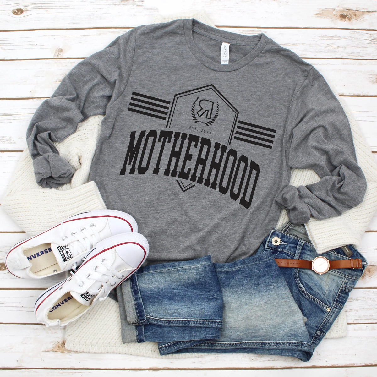 The Motherhood Collegiate Long Sleeved tee in gray triblend with a logo badge and unisex sizing is styled on a white wooden surface, paired with denim shorts, a white knit sweater, white Converse sneakers, and a brown watch.
